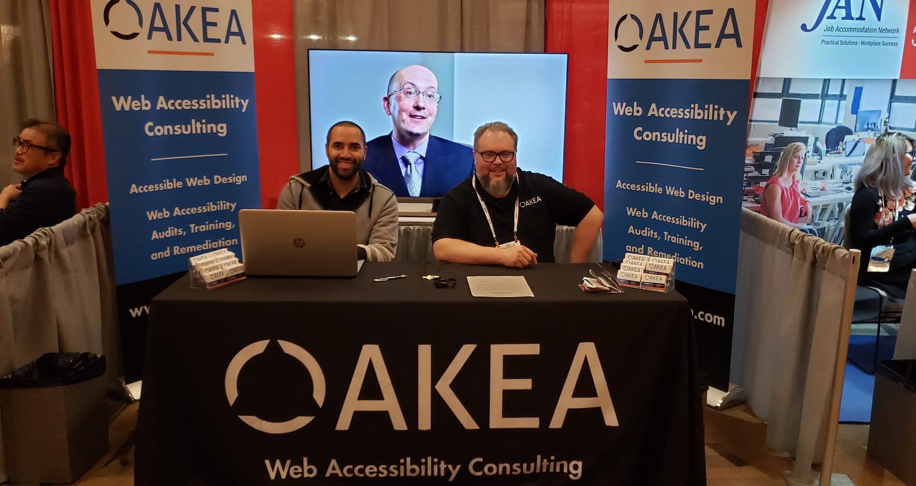 Ash Harris and a colleague at the AKEA Web Solutions booth at the CSUN Assistive Technology Conference, 2019, with a web accessibility segment playing on a screen behind them.