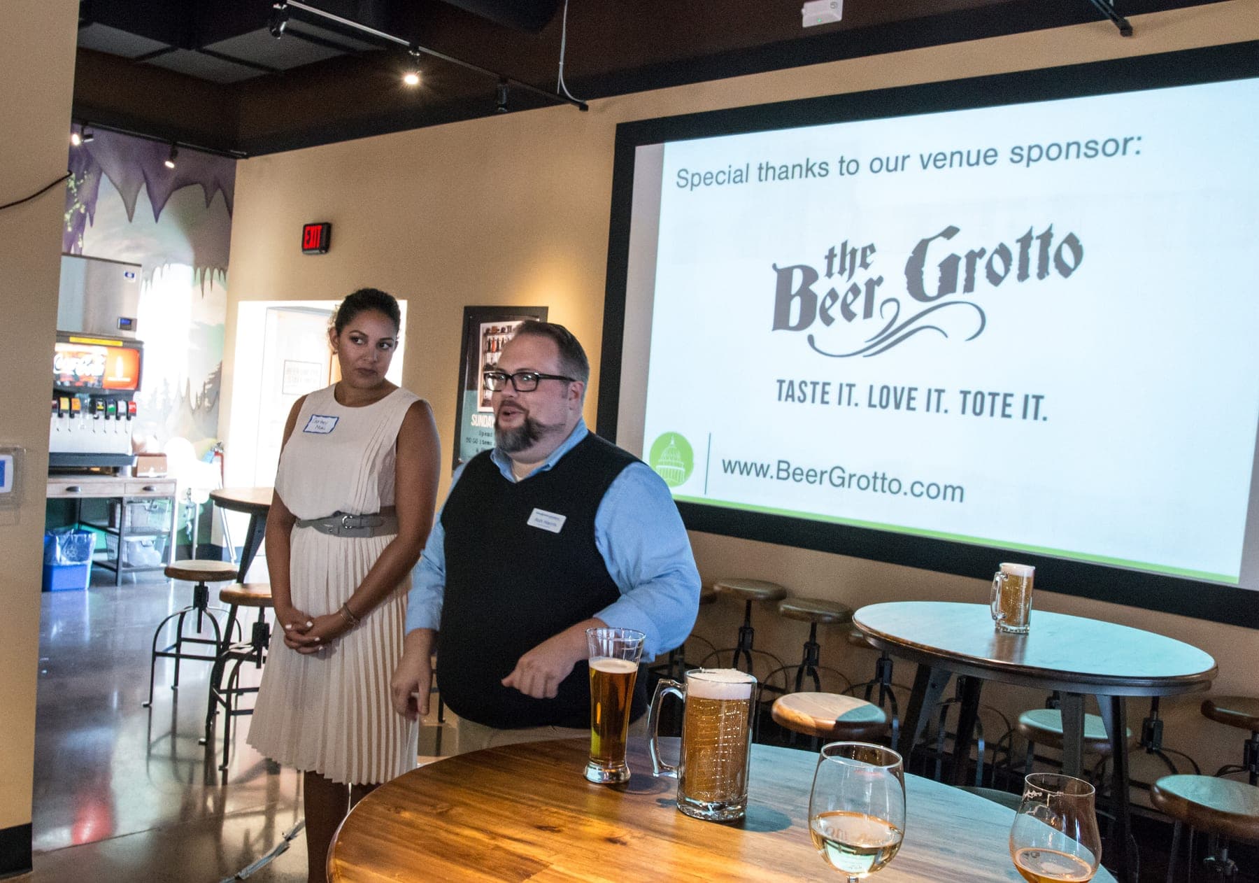 Ash Harris speaking to attendees at a Drinking Lunch networking event in Lansing, Michigan, a co-host beside him and the event's venue-sponsor slide projected on the screen behind.