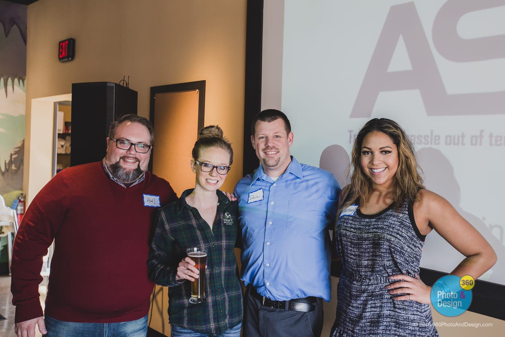 Ash Harris smiling with three other attendees at a Drinking Lunch event, name tags visible, beer in hand, projection screen in background.